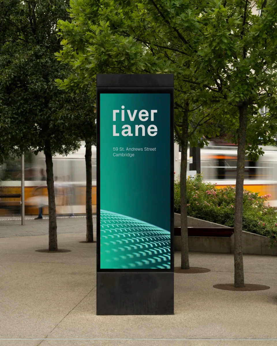 Outdoor digital sign displaying Riverlane branding and Cambridge office address at 59 St. Andrews Street, with trees and blurred tram in background.