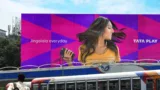 Image showing a Tata Play advert, featuring a woman listening to music and with the tagline 'Jingalala everyday'. Public transport is visible in the foreground, and buildings are in the far background.