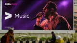 Image showing a billboard of a male performer, advertising the Tata Play music service. People are visible in the foreground, and there's a glimpse of buildings in the background.
